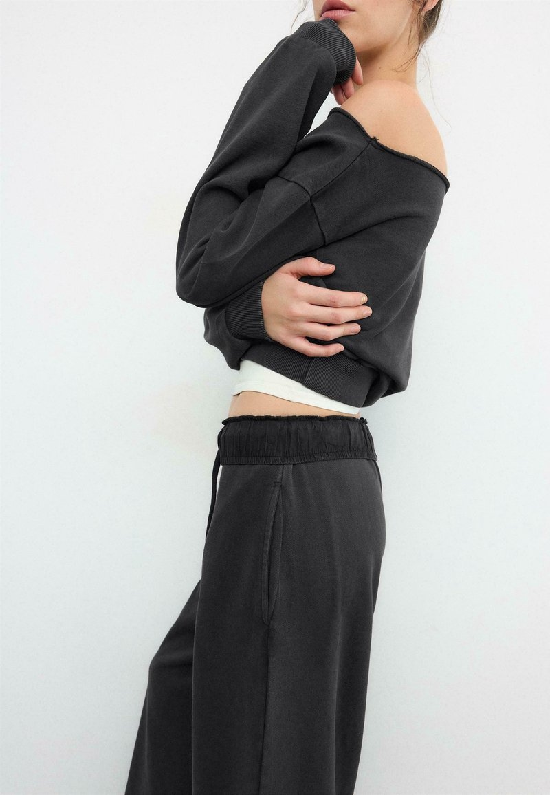 Femme en sweat-shirt noir épaules dénudées et pantalon noir assorti, bras croisés, sur fond blanc uni.