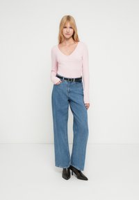 Ξανθιά γυναίκα στέκεται μπροστά σε λευκό φόντο φορώντας ανοιχτό ροζ μακρυμάνικο τοπ, τζιν παντελόνι με ψηλή μέση και φαρδιά μπατζάκια σε μπλε χρώμα, μαύρη ζώνη και μαύρα ψηλοτάκουνα μποτάκια.