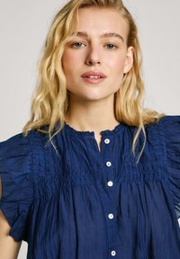 Blusa blu navy con corpino arricciato, maniche corte con volant e chiusura frontale con bottoni. Tessuto leggero e strutturato con una leggera lucentezza.