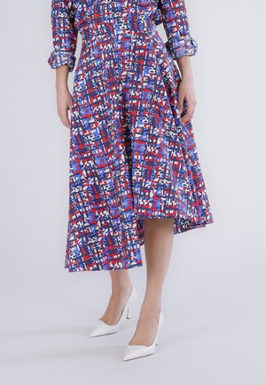 Kvinde iført en mellemlang kjole med blå, røde og hvide abstrakte mønstre og hvide stiletter, stående mod en ensfarvet baggrund.