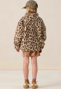 Kind dat met de rug naar de camera staat, gekleed in een roze met luipaardprint fuzzy jasje, bijpassende pet, lichtbruine rok, paarse sokken en beige sneakers.