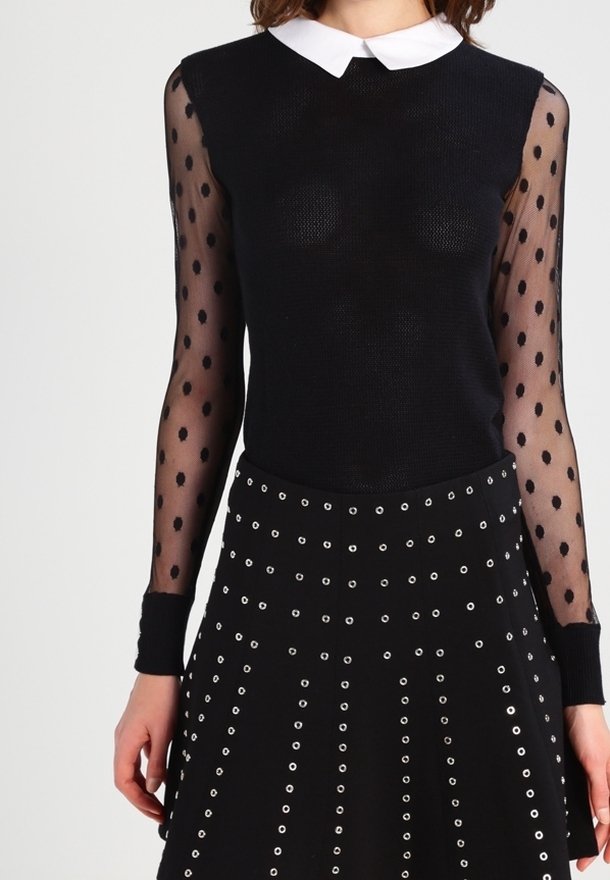 Femme portant un haut noir avec des manches longues transparentes à pois et un col blanc, associée à une jupe noire décorée d'œillets argentés.