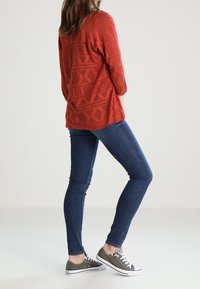 Persona vistiendo un suéter de patrón naranja óxido, jeans skinny azules y zapatillas grises, de pie con las manos en los bolsillos sobre un fondo sencillo.