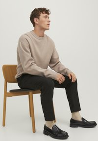 Pullover beige in maglia con scollo rotondo, pantaloni neri con texture, e mocassini neri abbinati a calzini beige, seduto su una sedia di legno.