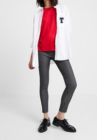 Mujer vistiendo una camisa blanca sobre una blusa roja, pantalones ajustados negros de rayas y zapatos de cordones negros brillantes, de pie contra un fondo blanco.