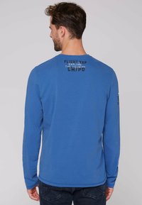 Niebieska bawełniana koszulka z długim rękawem i okrągłym dekoltem, z grafikami w czerni i szarości na górnej części pleców oraz lewym rękawie.