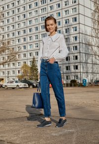 Camisa branca de botão com um grande colarinho, jeans azuis de cintura alta, ténis azul-marinho e uma bolsa de mão a condizer, com um fundo de um edifício moderno.