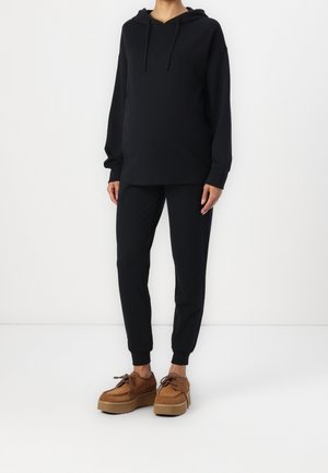 Personne portant un sweat à capuche noir et un pantalon de jogging noir avec des chaussures compensées beige, debout devant un fond blanc uni.