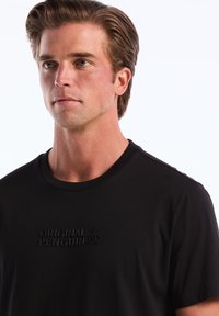 T-shirt en coton noir avec un logo "Original Penguin" texturé sur la poitrine. Il possède un col rond et des manches courtes.