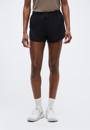 Άτομο που φοράει μαύρο αθλητικό σορτς, μπεζ μπλούζα, λευκές κάλτσες και άσπρα αθλητικά παπούτσια, στέκεται μπροστά σε ένα απλό φωτεινό φόντο.