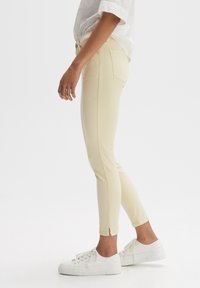 Pantalon jaune coupe slim avec une texture lisse et des fentes aux chevilles, porté avec des baskets blanches. Vue de côté mettant en valeur la coupe et le style.