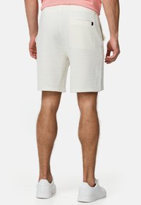 Pantalones cortos blancos texturizados con bolsillos laterales y un bolsillo trasero, combinados con zapatillas blancas. El diseño es simple y llega hasta la rodilla.