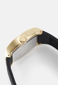 Caja de reloj rectangular en tono dorado con correa de silicona negra. Presenta un acento texturizado en la correa y una corona en el lateral para ajustes.