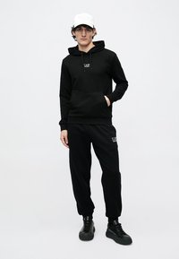 Μαύρο φούτερ με κουκούλα, μπροστινή τσέπη και λογότυπο στο στήθος, συνδυασμένο με μαύρο παντελόνι φόρμας, φορεμένο με λευκό καπέλο και μαύρα αθλητικά παπούτσια.