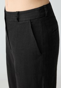 Schwarze Leinenhose mit geradem Schnitt, ausgestattet mit einem Bund, vorderen Falten und einer Seitentasche für praktische Nutzung. Glatte Textur.