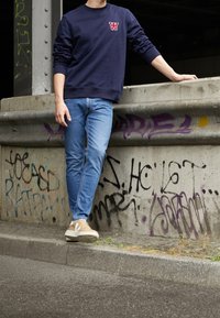Mörkblå sweatshirt med röd logotyp, blå jeans och beige sneakers med vita detaljer. Betongbakgrund med graffitivägg och trottoar.