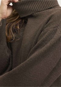 Kvinna med långt hår som bär en brun ribbad polotröja och håller kragen nära sitt ansikte med ena handen.