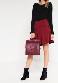 Batoh z bordové kůže s horním klopou, kovovým kroužkem a dvěma rukojeťmi. Texturovaný povrch s čistým, obdélníkovým tvarem.