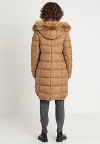 Abrigo marrón acolchado con capucha de piel, con un diseño acolchado y corte a media longitud, combinado con pantalones grises y botas negras.