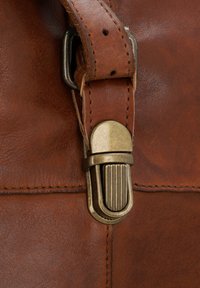 Close-up of a brass metal clasp attached to a brown leather strap on a stitched brown leather bag.