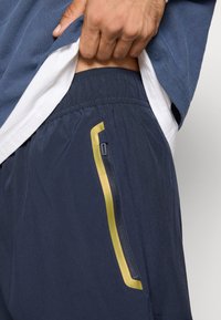 Μπλε σορτς με υφασμάτινη υφή, ελαστική μέση και έντονη χρυσή λεπτομέρεια κατά μήκος της φερμουάρ πλευρικής τσέπης.