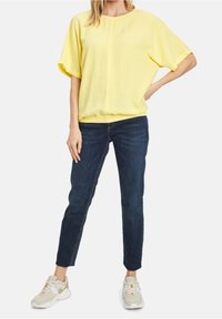Żółta luźna bluzka z krótkimi rękawami i ściąganym dołem, połączona z ciemnoniebieskimi obcisłymi dżinsami i jasnymi trampkami.