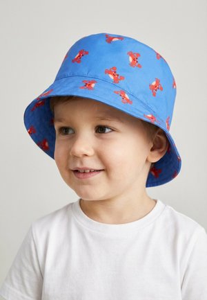 Junger Junge lächelt, trägt einen blauen Fischerhut mit roten Krabbenmotiven und ein weißes T-Shirt, leicht nach rechts gewandt.