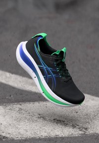 Chaussure de sport noire avec des accents verts et bleus, design léger, semelle rembourrée et dessus texturé, dotée d'un système de laçage.