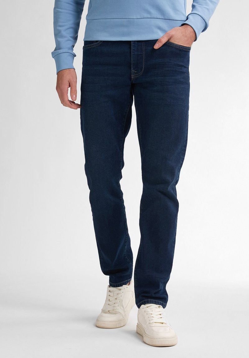 Vaqueros de mezclilla azul oscuro con corte ajustado, que cuentan con bolsillos frontales y pierna recta, combinados con zapatillas blancas. Textura suave.