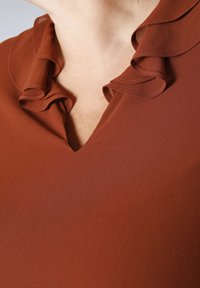 Close-up van een roestkleurige top met een gerimpelde V-halskraag op de bovenste borst en nek van een persoon.