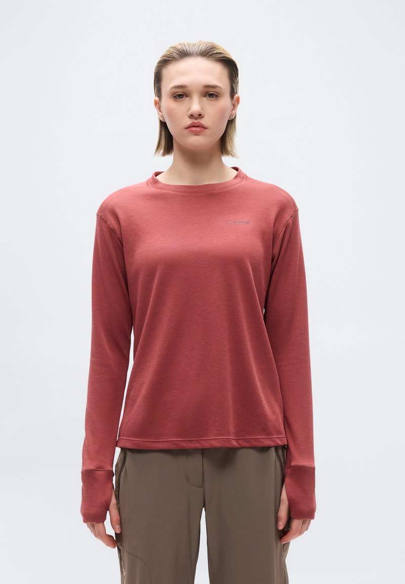 Noor inimene, kes vaatab otse, kannab roostes värvi pika varrukaga tippu pöidla aukudega ja oliivirohelisi cargo-pükse, taustaks on ühtlane värv.