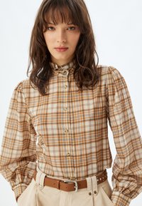 Bruine en crèmekleurige geruite blouse met lange pofmouwen en een gerimpelde kraag, voorzien van gouden knopen en een bruine riem met studs.