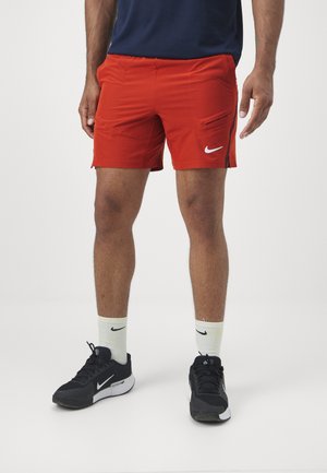 Mężczyzna w jaskrawoczerwonych sportowych szortach Nike, białych skarpetkach Nike do kostek i czarnych butach do biegania Nike, stojący na jednolitym białym tle.
