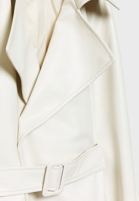 Gros plan d’un trench-coat crème montrant de larges revers et une ceinture assortie avec une boucle rectangulaire.