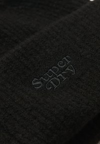 Gorro de punto negro con una tela texturizada. Presenta un logo bordado de "Super Dry" en hilo oscuro. En general, un diseño simple y discreto.
