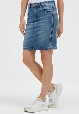 Frau trägt knielangen blauen Jeansrock mit seitlichem Reißverschluss und weißen Sneakers, steht vor weißem Hintergrund.