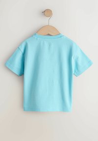 Svetlomodré bavlnené tričko s krátkym rukávom zavesené na drevenom ramienku. Má okrúhly výstrih a jednoduchý, čistý dizajn.