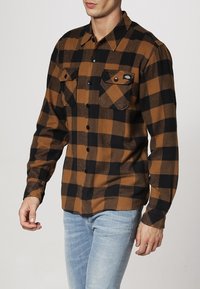 Brun och svart rutigt flanellskjorta med knäppning fram, två bröstfickor och krage. Har en mjuk textur och långa ärmar.