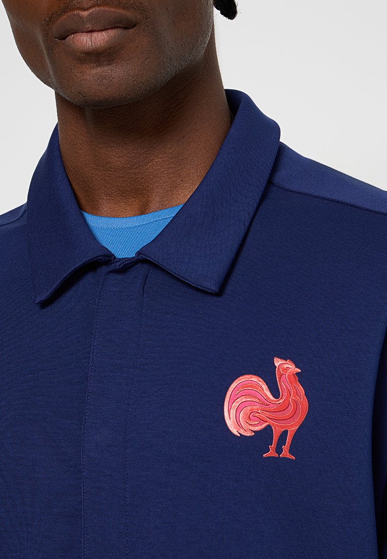Marineblaues Poloshirt mit strukturierter Kragen und einem lebhaften pinkfarbenen, gestickten Hahndetail auf der linken Brust. Blause Unterhemd darunter sichtbar.