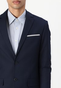 Navy blazer with notched lapels, two buttons, and side pockets, paired with a light blue collared shirt and a silver pocket square.