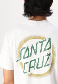 Bijela pamučna majica s grafikom u zelenoj i zlatnoj boji u obliku kruga na leđima, s natpisom "SANTA CRUZ" u podebljanom, istrošenom fontu.