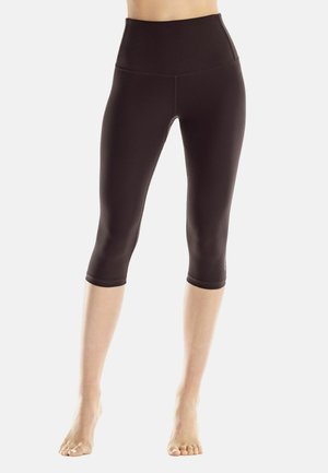 Frau trägt schwarze, hoch taillierte, dreiviertellange enge Leggings und steht barfuß auf hellem, schlichtem Hintergrund.