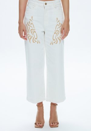 Pantalon blanc à jambes larges avec motifs de flammes brodés beige sur les cuisses, porté avec des talons beiges à bout ouvert et bride à la cheville sur fond blanc.