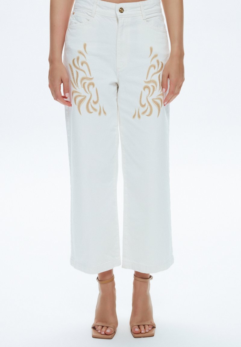 Pantalon blanc à jambes larges avec motifs de flammes brodés beige sur les cuisses, porté avec des talons beiges à bout ouvert et bride à la cheville sur fond blanc.