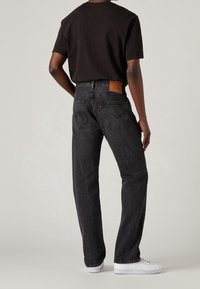 Jeans de mezclilla negra con corte recto, que cuentan con un parche de cuero marrón en la parte trasera de la cinturilla y bolsillos traseros clásicos.