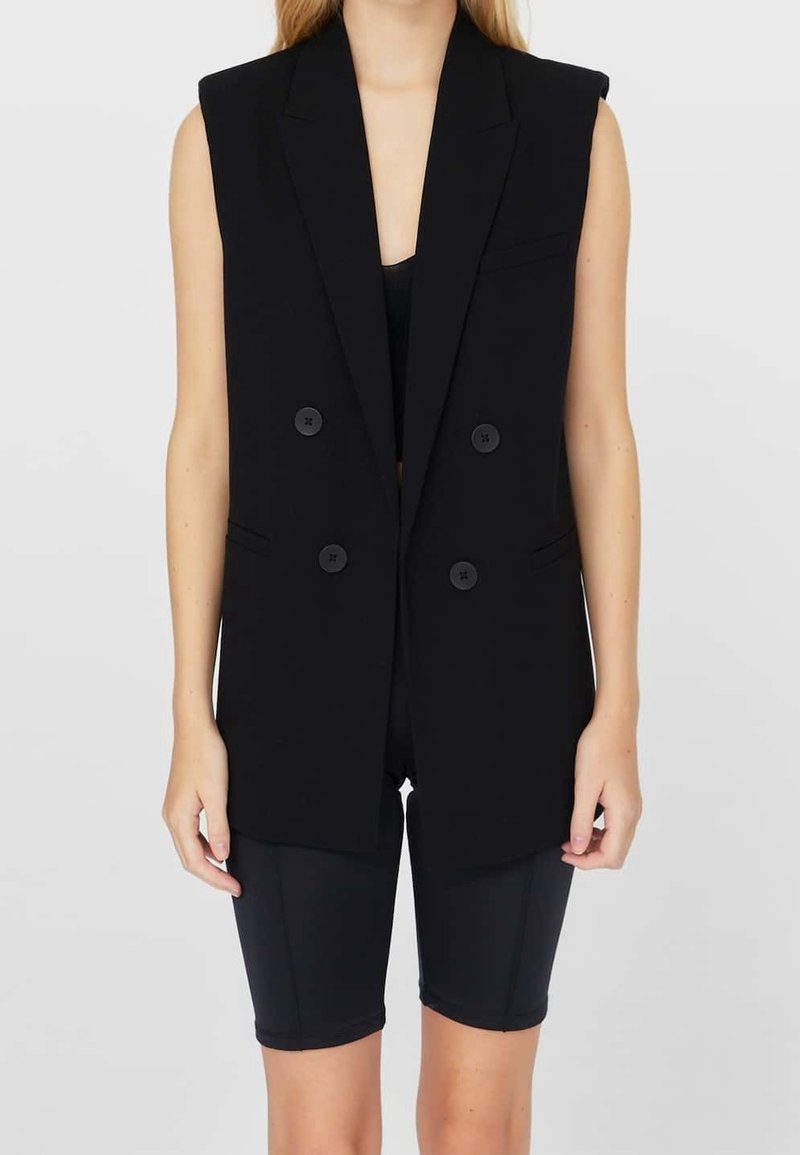 Black sleeveless blazer with a double-breasted design, featuring large buttons and a pocket. Worn over black cycling shorts.