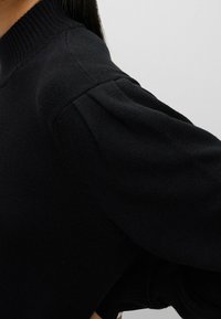 Pull noir en maille avec un col montant côtelé et des manches légèrement bouffantes. La texture est douce, avec une surface lisse et une coupe ajustée.