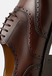 Braune Lederdressschuhe mit Broguedetails, runder Zehenform und dünnen Schnürsenkeln. Glatte Textur mit glänzendem Finish und gestapeltem Absatz.