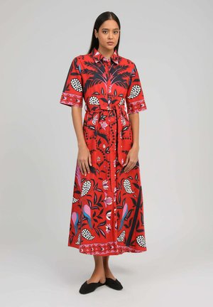Femme debout portant une robe maxi rouge boutonnée avec ceinture, ornée de motifs floraux et d'oiseaux noirs, blancs, roses et bleus, associée à des chaussures slip-on noires.