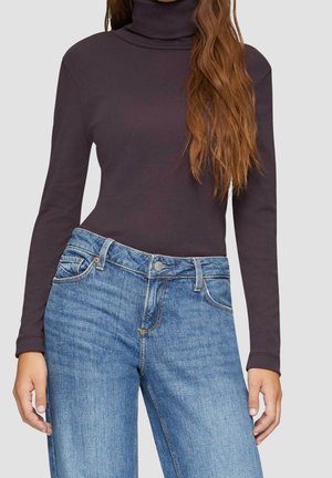 Vrouw draagt een nauwsluitende donkerpaarse coltrui en hooggetailleerde blauwe spijkerbroek met lang golvend bruin haar over één schouder.